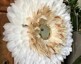 May include: A white and brown feather wreath with a round mirror in the centre. The wreath is hanging on a wooden cabinet.
