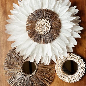 Op de afbeelding: Drie ronde wanddecoraties in boho-chique stijl. Het grootste stuk is een witte verenkrans met een bruin en wit schelp-centrum. De twee kleinere stukken zijn gemaakt van natuurlijke materialen, een met een bruin geweven centrum en de andere met een wit schelp-centrum.