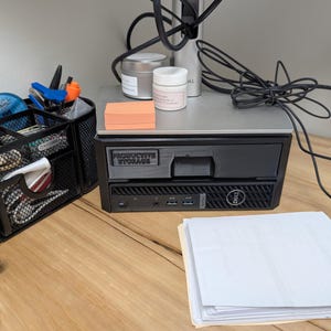 May include: A black desktop computer with the text "PRODUCTIVE STORAGE" is on a wooden desk. A black mesh organizer holds pens, tape, and other office supplies. A stack of orange sticky notes and a few jars are on the computer.