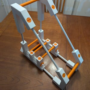 May include: A white and orange adjustable stand featuring a rectangular frame with multiple horizontal orange bars, vertical white supports, and orange circular screw-like fasteners connecting the structural components of the device.