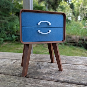 May include: A miniature blue nightstand with two drawers and white curved handles. The nightstand has a brown frame and sits on four brown legs. The piece is displayed on a weathered wooden surface.