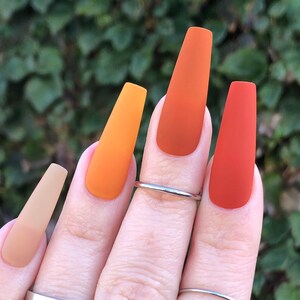 May include: Close-up of a hand with long, almond-shaped nails painted in a gradient of orange, yellow, and brown shades. The nails are matte and have a smooth finish.