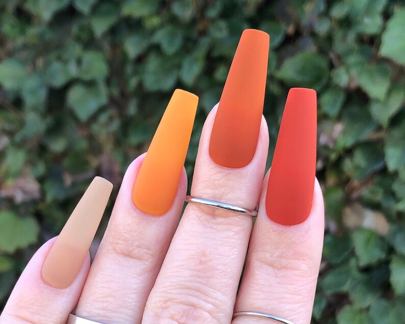May include: Close-up of a hand with long, almond-shaped nails painted in a gradient of orange, yellow, and brown shades. The nails are matte and have a smooth finish.