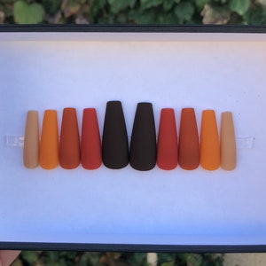 May include: A set of ten false nails in various shades of brown, orange, and beige. The nails are arranged in a row in a black box.