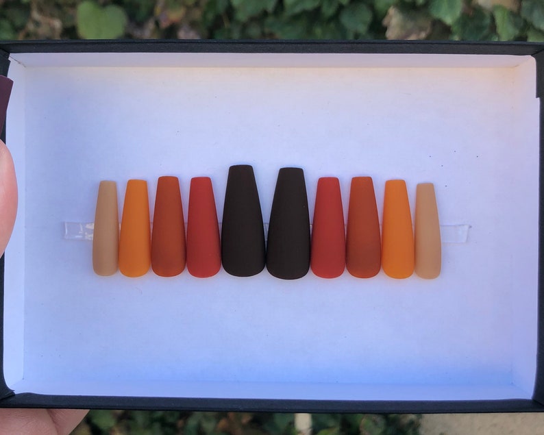 May include: A set of ten false nails in various shades of brown, orange, and beige. The nails are arranged in a row in a black box.