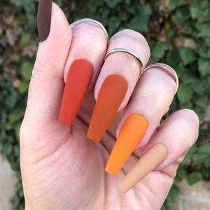 May include: A set of long, almond-shaped false nails with a gradient of orange, brown, and beige colors. The nails have a matte finish.
