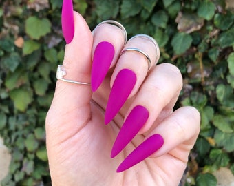 May include: A hand with long, stiletto-shaped nails painted a vibrant magenta color. The nails have a matte finish.