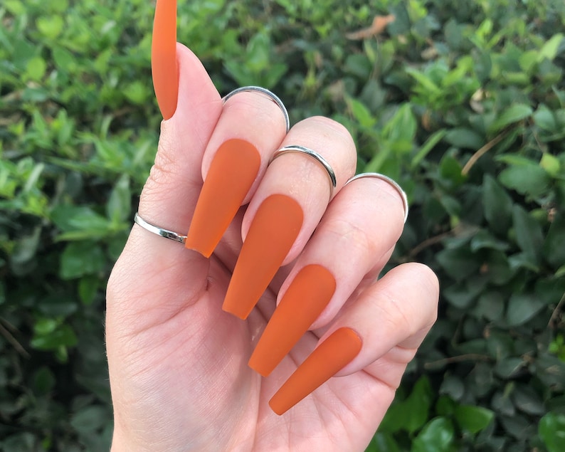 May include: Long, almond-shaped, matte orange press-on nails.