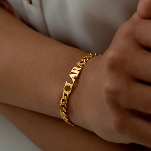 Op de afbeelding: Een goudkleurige armband met de naam "CLARA" in hoofdletters. De armband heeft een gourmette ketting ontwerp. De naam is geïntegreerd in de schakels van de ketting, waardoor een gepersonaliseerd accessoire ontstaat.