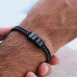 Könnte beinhalten: Ein schwarzes Kettenarmband mit vier kleinen Würfeln. Jeder Würfel trägt einen Namen in Weiß: Luca, Emma, Noah und Eva. Das Armband wird am Handgelenk getragen.