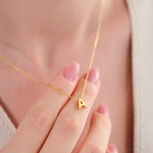 Peut inclure: Un collier doré délicat avec un petit pendentif en forme de lettre "A". Le pendentif est suspendu à une fine chaîne. Le collier est présenté sur le cou et la main d'une personne, avec un fond neutre.