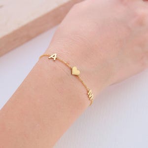 May include: A delicate gold bracelet with the letters "A" and "M" and a heart charm. The bracelet is shown on a wrist, highlighting its fine chain and minimalist design. The bracelet is a piece of jewellery.
