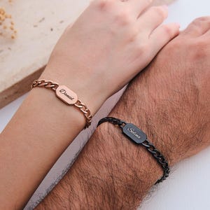 K&ouml;nnte beinhalten: Zwei personalisierte Armb&auml;nder mit Kettengliedern. Eines ist ros&eacute;goldfarben mit dem Namen "Daniel" auf einer rechteckigen Platte eingraviert. Das andere ist schwarz mit dem Namen "Selina" auf einer &auml;hnlichen Platte eingraviert. Die Armb&auml;nder werden an Handgelenken getragen.