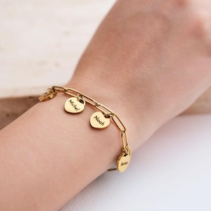 Puede incluir: Pulsera dorada con diseño de eslabones de cadena. La pulsera presenta tres colgantes en forma de corazón, cada uno grabado con un nombre: "Michel", "Noah" y "Anna". La pulsera se lleva en la muñeca.