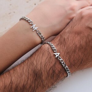 Op de afbeelding: Twee zilveren kettingarmbanden, elk met een enkele initiaal. Eén armband toont de letter "M", de andere de letter "A". De armbanden zitten om polsen, wat duidt op bijpassende sieraden voor een stel.