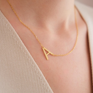 Peut inclure: Un collier doré avec la lettre majuscule "A" en guise de pendentif. La lettre est reliée à une chaîne délicate. Le collier est porté autour du cou, le pendentif reposant près de la clavicule.