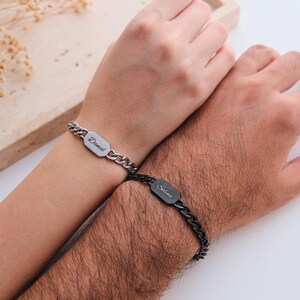 K&ouml;nnte beinhalten: Zwei passende Kettenarmb&auml;nder, eines silberfarben und eines schwarz, jeweils mit einem rechteckigen Namensschild. Auf dem silbernen Armband steht "Daniel", auf dem schwarzen Armband "Selina". Die Armb&auml;nder werden an den Handgelenken getragen.