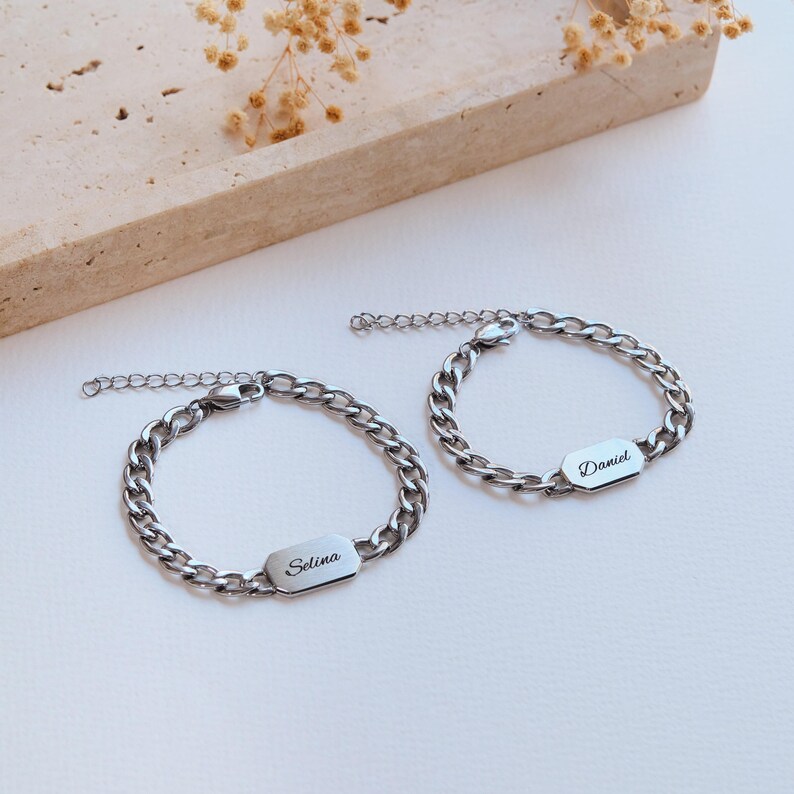 K&ouml;nnte beinhalten: Zwei silberfarbene Kettenarmb&auml;nder mit rechteckigen Namensschildern. Eines ist mit "Selina" graviert, das andere mit "Daniel". Die Armb&auml;nder liegen auf einer wei&szlig;en Oberfl&auml;che mit einem beigen Dekorationselement im Hintergrund.