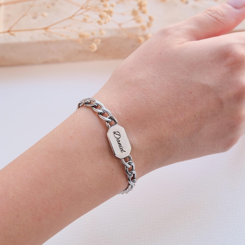 K&ouml;nnte beinhalten: Silberfarbenes Armband mit Kettengliedern und einem rechteckigen Namensschild. Auf dem Schild steht der Name "Daniel" in Schreibschrift. Das Armband wird am Handgelenk getragen.