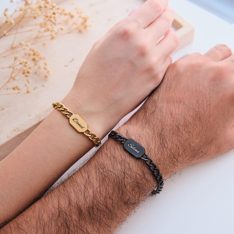 K&ouml;nnte beinhalten: Zwei passende Armb&auml;nder, eines goldfarben und eines schwarz, jeweils mit einem rechteckigen Namensschild. Auf dem goldenen Armband steht "Daniel" und auf dem schwarzen Armband steht "Selina". Beide Armb&auml;nder haben ein Panzerketten-Design.