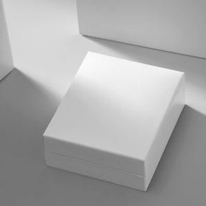 K&ouml;nnte beinhalten: Eine wei&szlig;e quadratische Box mit Deckel, die auf einer grauen Oberfl&auml;che steht. Die Box ist scharf, der Hintergrund unscharf.