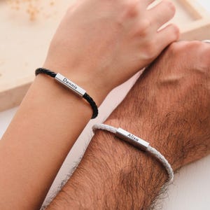 Puede incluir: Dos pulseras de cuero trenzado, una negra y otra blanca, cada una con una barra plateada grabada con un nombre. La pulsera negra dice "Dennis", y la pulsera blanca dice "Alisa". Las pulseras se llevan en las muñecas.