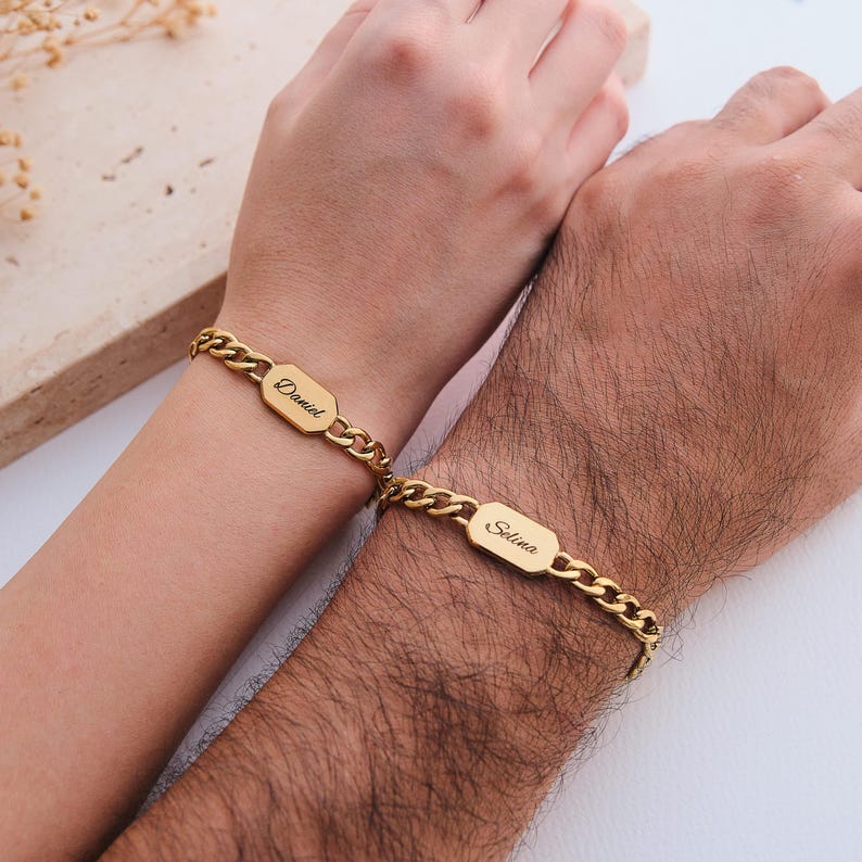 K&ouml;nnte beinhalten: Goldfarbene Kettenarmb&auml;nder, jeweils mit einem rechteckigen Anh&auml;nger mit eingravierten Namen. Auf einem Armband steht "Daniel", auf dem anderen "Selina". Die Armb&auml;nder werden an Handgelenken getragen und zeigen ein passendes Schmuckset.
