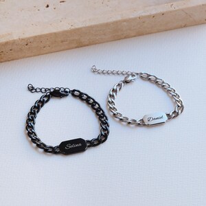 K&ouml;nnte beinhalten: Zwei personalisierte Kettenarmb&auml;nder. Eines ist schwarz mit "Selina" graviert auf einer rechteckigen Platte. Das andere ist silberfarben mit "Daniel" graviert. Beide Armb&auml;nder haben verstellbare Verschl&uuml;sse und liegen auf einer wei&szlig;en Oberfl&auml;che.