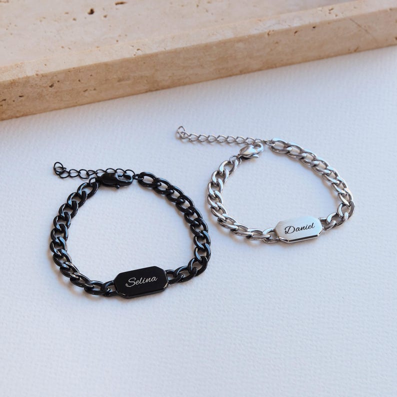 K&ouml;nnte beinhalten: Zwei personalisierte Kettenarmb&auml;nder. Eines ist schwarz mit "Selina" graviert auf einer rechteckigen Platte. Das andere ist silberfarben mit "Daniel" graviert. Beide Armb&auml;nder haben verstellbare Verschl&uuml;sse und liegen auf einer wei&szlig;en Oberfl&auml;che.