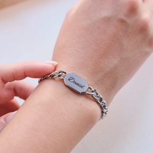 K&ouml;nnte beinhalten: Ein silberfarbenes Armband mit Kettenglied-Design. Das Armband hat eine rechteckige Platte mit dem eingravierten Namen "Daniel". Das Armband wird am Handgelenk getragen.