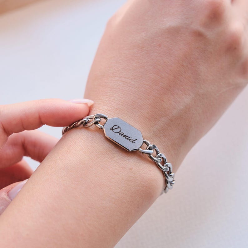 K&ouml;nnte beinhalten: Ein silberfarbenes Armband mit Kettenglied-Design. Das Armband hat eine rechteckige Platte mit dem eingravierten Namen "Daniel". Das Armband wird am Handgelenk getragen.