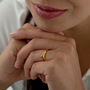 May include: A gold-toned ring with the name "Selena" spelled out in cursive script.