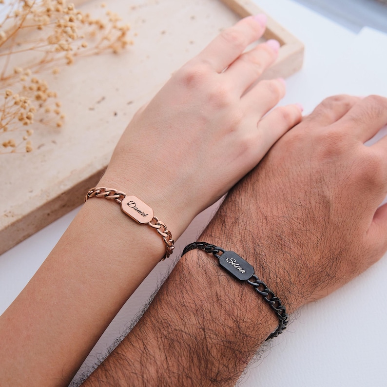 K&ouml;nnte beinhalten: Zwei personalisierte Armb&auml;nder mit Kettengliedern. Eines ist ros&eacute;goldfarben mit dem Namen "Daniel" auf einer rechteckigen Platte eingraviert. Das andere ist schwarz mit dem Namen "Selina" auf einer &auml;hnlichen Platte eingraviert. Die Armb&auml;nder werden an Handgelenken getragen.