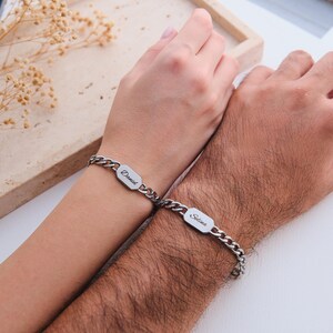 K&ouml;nnte beinhalten: Zwei silberne Kettenarmb&auml;nder, jeweils mit einem rechteckigen Namensschild. Auf einem Armband steht "Daniel" und auf dem anderen "Selina". Die Armb&auml;nder befinden sich an Handgelenken, wobei die H&auml;nde zusammengelegt sind.