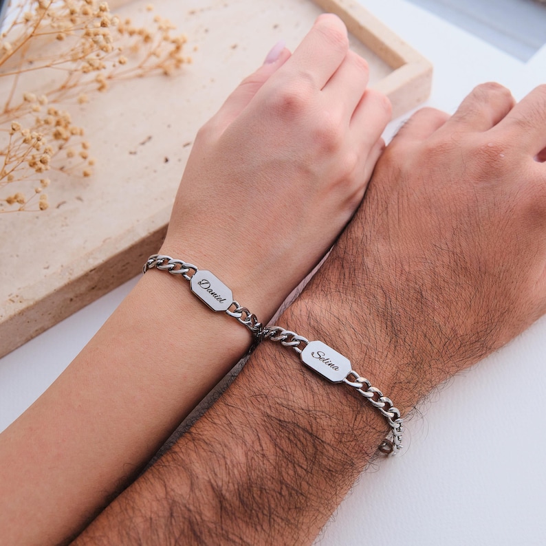 K&ouml;nnte beinhalten: Zwei silberne Kettenarmb&auml;nder, jeweils mit einem rechteckigen Namensschild. Auf einem Armband steht "Daniel" und auf dem anderen "Selina". Die Armb&auml;nder befinden sich an Handgelenken, wobei die H&auml;nde zusammengelegt sind.