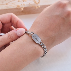 K&ouml;nnte beinhalten: Ein silberfarbenes Kettenarmband mit einem rechteckigen Namensschild mit dem eingravierten Namen "Daniel". Das Armband befindet sich an einem Handgelenk. Die Kettenglieder sind dick und haben eine abgerundete Form.