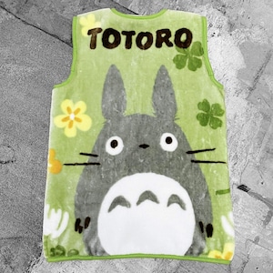 May include: A green sleeveless apron with the word "TOTORO" at the top, featuring the character Totoro from the animated film My Neighbour Totoro. The apron has a large illustration of Totoro, with yellow flowers and green clovers.
