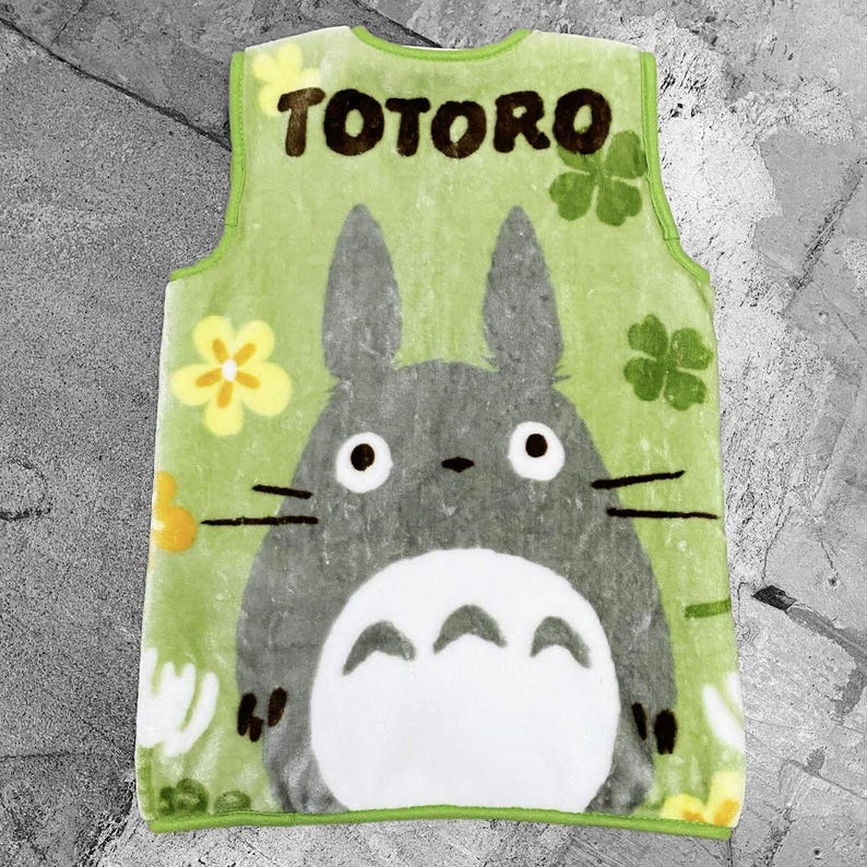May include: A green sleeveless apron with the word "TOTORO" at the top, featuring the character Totoro from the animated film My Neighbour Totoro. The apron has a large illustration of Totoro, with yellow flowers and green clovers.