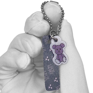 May include: A keychain featuring a silver ball chain and a purple teddy bear charm. The charm is connected to a dark blue rectangular tag with white text and small white crosses.