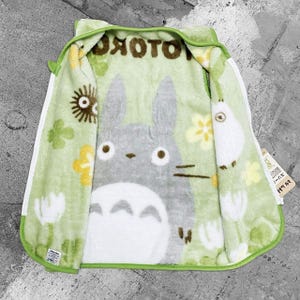 May include: A green hooded zip-up blanket with a large gray Totoro character on the inside. The blanket has a white zip and trim, with a light green exterior and a pattern of flowers and other characters.