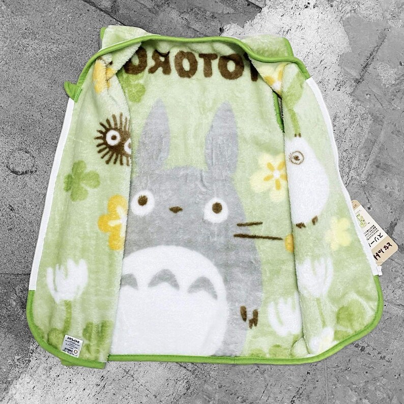 May include: A green hooded zip-up blanket with a large gray Totoro character on the inside. The blanket has a white zip and trim, with a light green exterior and a pattern of flowers and other characters.