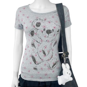 May include: A gray t-shirt with a round neckline featuring a cartoon bear design and pink floral accents. The text "Charming Bear" and "Love me & Love you" are printed on the shirt. A white bunny charm hangs from a black denim bag.