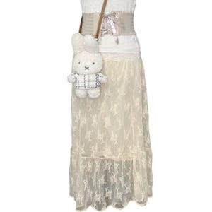 May include: A white lace skirt with a ruffled hem and a white plush bunny bag with a plaid shirt. The skirt is worn with a white top and a brown corset belt.