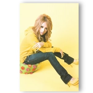 Puede incluir: Una mujer con una sudadera amarilla y pantalones vaqueros azules se sienta en un cojín redondo de colores sobre un fondo amarillo. Lleva tacones altos amarillos.