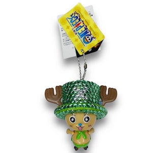 Puede incluir: Un llavero de One Piece con un personaje verde y marrón con brillantes. El personaje lleva un sombrero verde con cuernos y tiene una gran sonrisa. El llavero está unido a una cadena plateada.