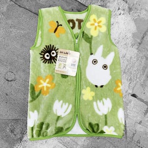 May include: A green sleep vest with a zip fastening. The vest is decorated with a cartoon design featuring a white rabbit-like character, yellow flowers, and a black, round creature. The vest has a tag with Japanese text.