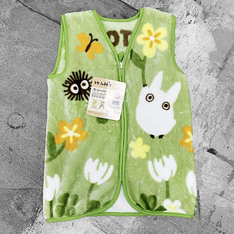 May include: A green sleep vest with a zip fastening. The vest is decorated with a cartoon design featuring a white rabbit-like character, yellow flowers, and a black, round creature. The vest has a tag with Japanese text.