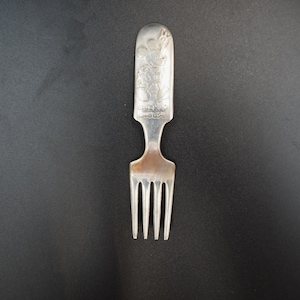 May include: A silver fork with a decorative handle featuring an embossed design. The handle has a rounded top and a slender neck leading to the four tines. The fork is set against a dark background.