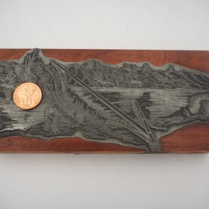 May include: A rectangular wooden printing block featuring a raised metal plate with an engraved landscape scene of a body of water and mountains. A copper-colored penny is placed on the left side of the metal plate for scale.