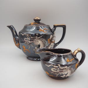 Peut inclure: Service à thé en céramique noire avec des détails dorés et des motifs de dragons. La théière a un couvercle et une anse, la crémière un bec et une anse. L'ensemble est pour servir le thé.